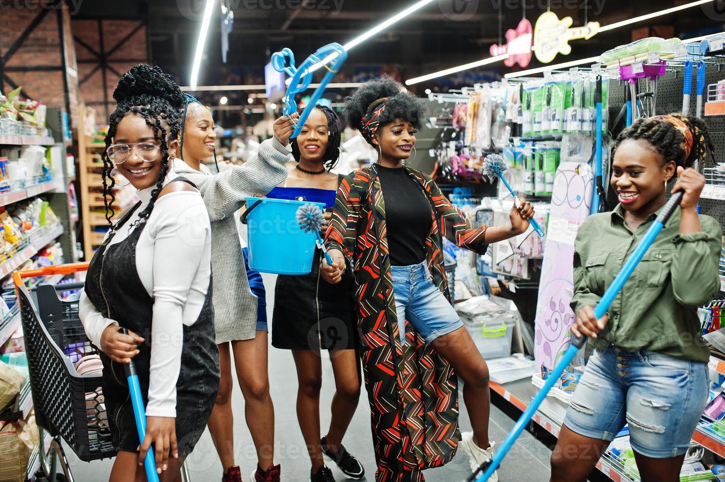 Group of five african womans with dust mop, toilet brush and bucket having fun in household cleaning items department in supermarket. photo