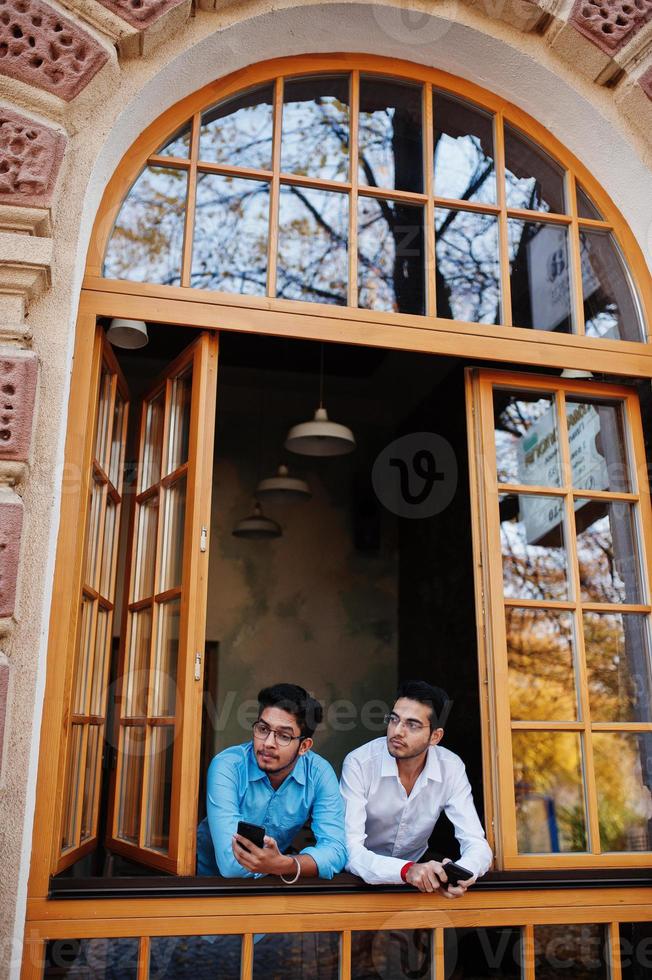 dos hombres del sur de asia posaron en una reunión de negocios en un café. los indios trabajan juntos usando varios aparatos, conversando. sosteniendo teléfonos móviles. foto