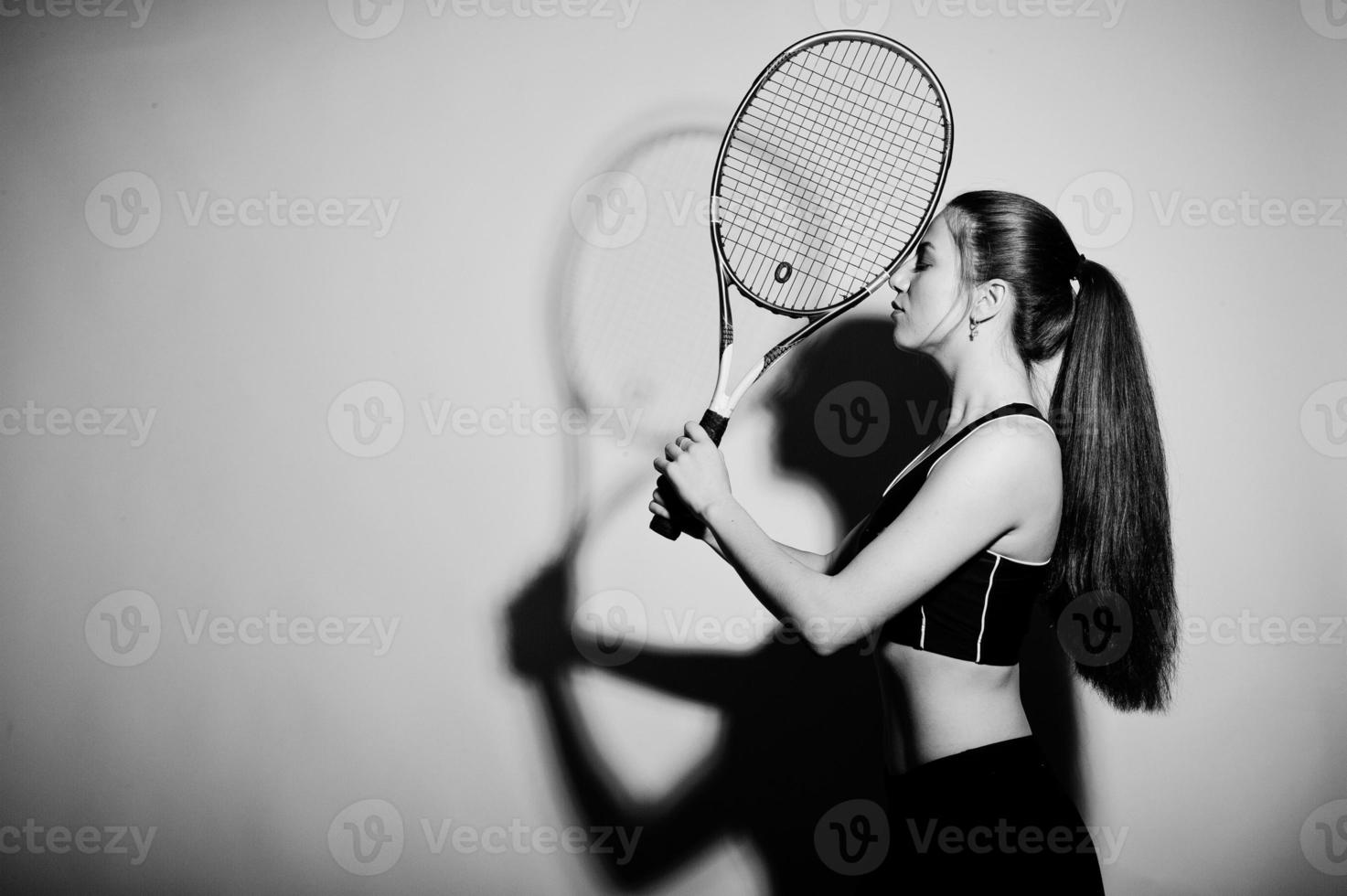 Black and white portrait of beautiful young woman player in sports clothes holding tennis racket while standing against white background. photo