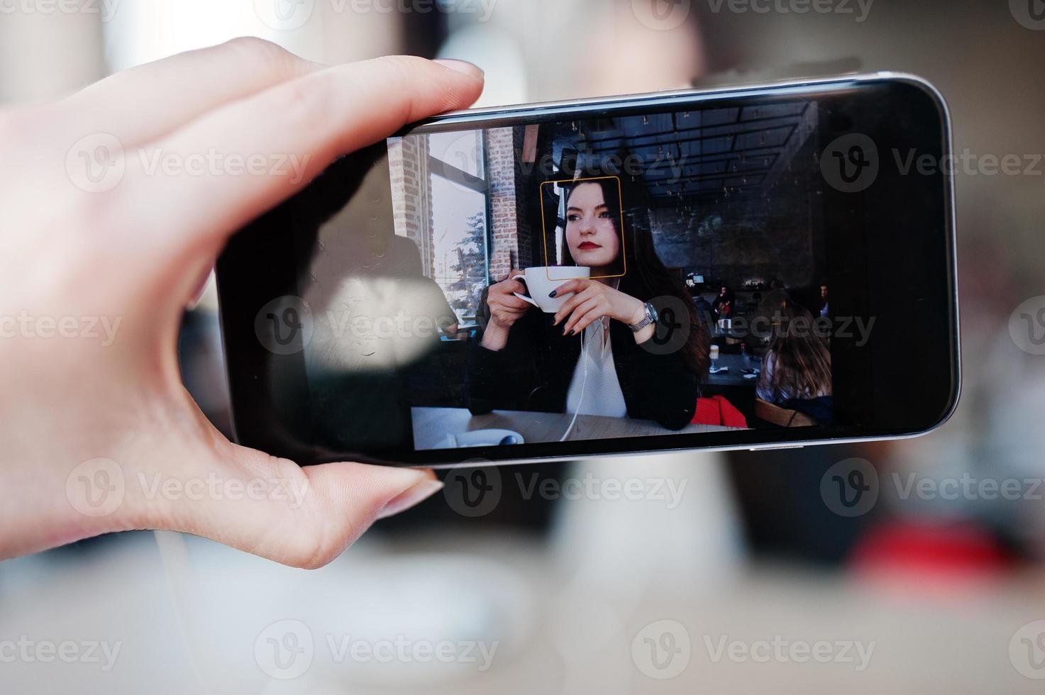 Close up photo of screen mobile phones with brunette girl sitting on cafe with cup of cappuccino, listening music on headphones.