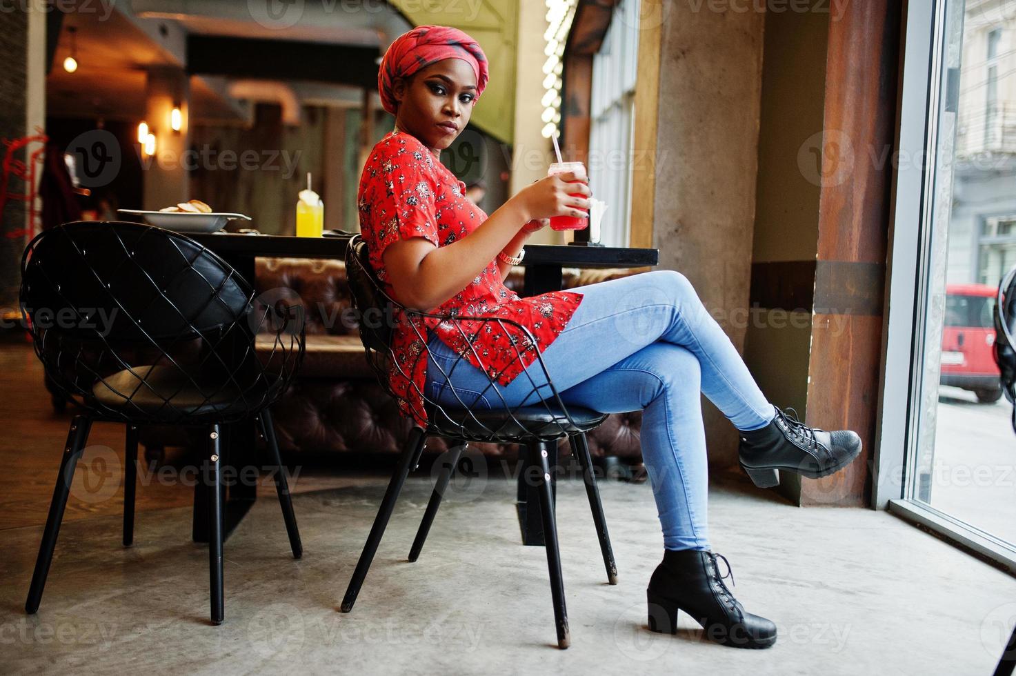 Stylish african woman in red shirt and hat posed indoor cafe and drinking strawberry lemonade with phone at hand. photo