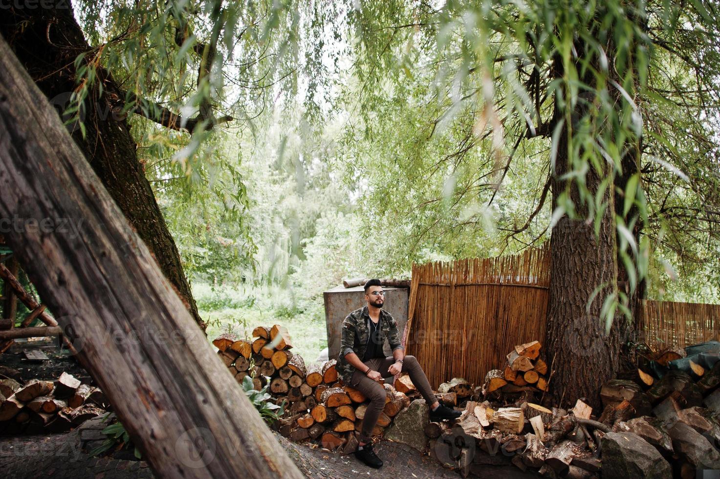 Impresionante y hermoso hombre macho alto de barba árabe con gafas y chaqueta militar posado al aire libre, sentado en tocones. foto