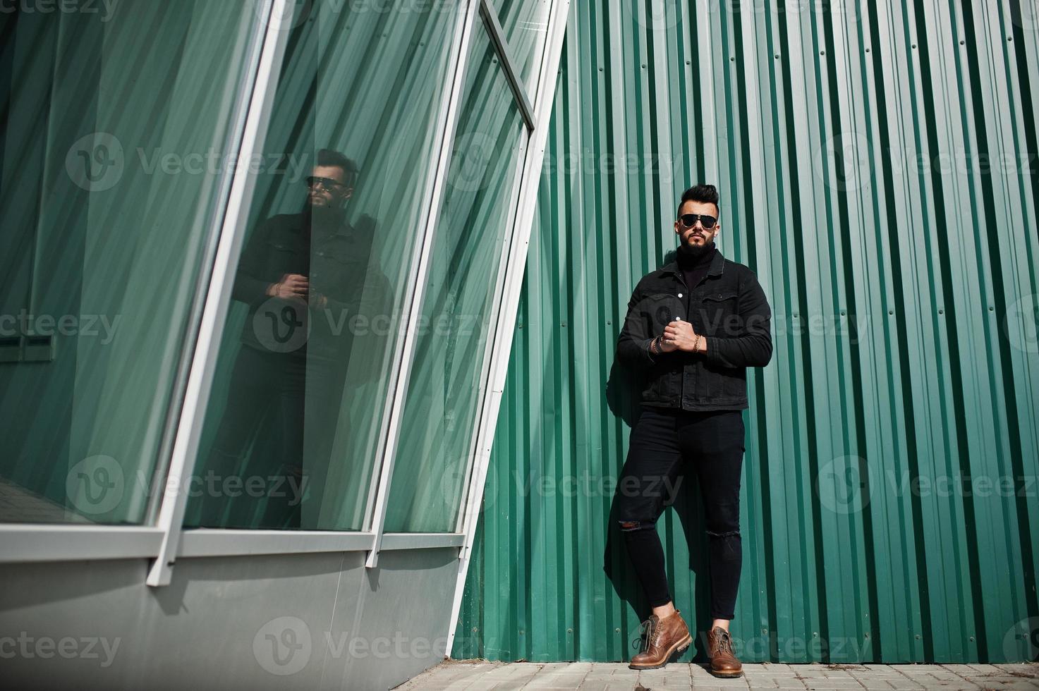 El hombre árabe de barba rica en moda viste una chaqueta negra de jeans y gafas de sol posadas contra una pared verde con ventanas. chico modelo árabe elegante, exitoso y de moda. foto