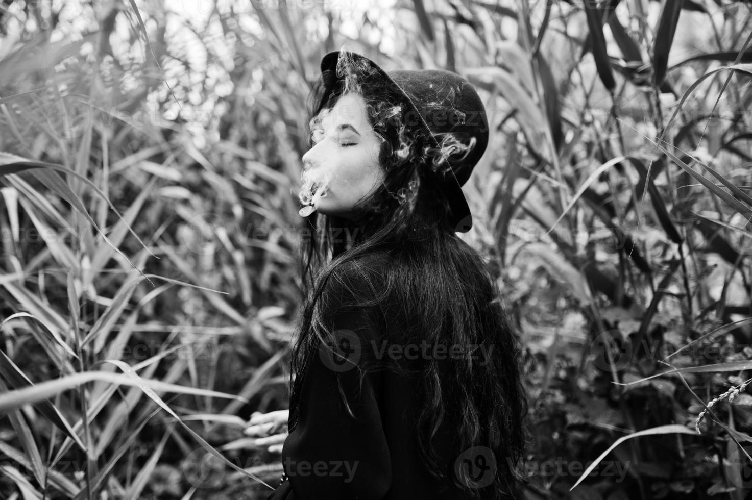 Sensual smoker girl all in black, red lips and hat. Goth dramatic woman smoking on common reed. Black and white portrait. photo