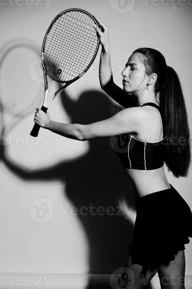Black and white portrait of beautiful young woman player in sports clothes holding tennis racket while standing against white background. photo
