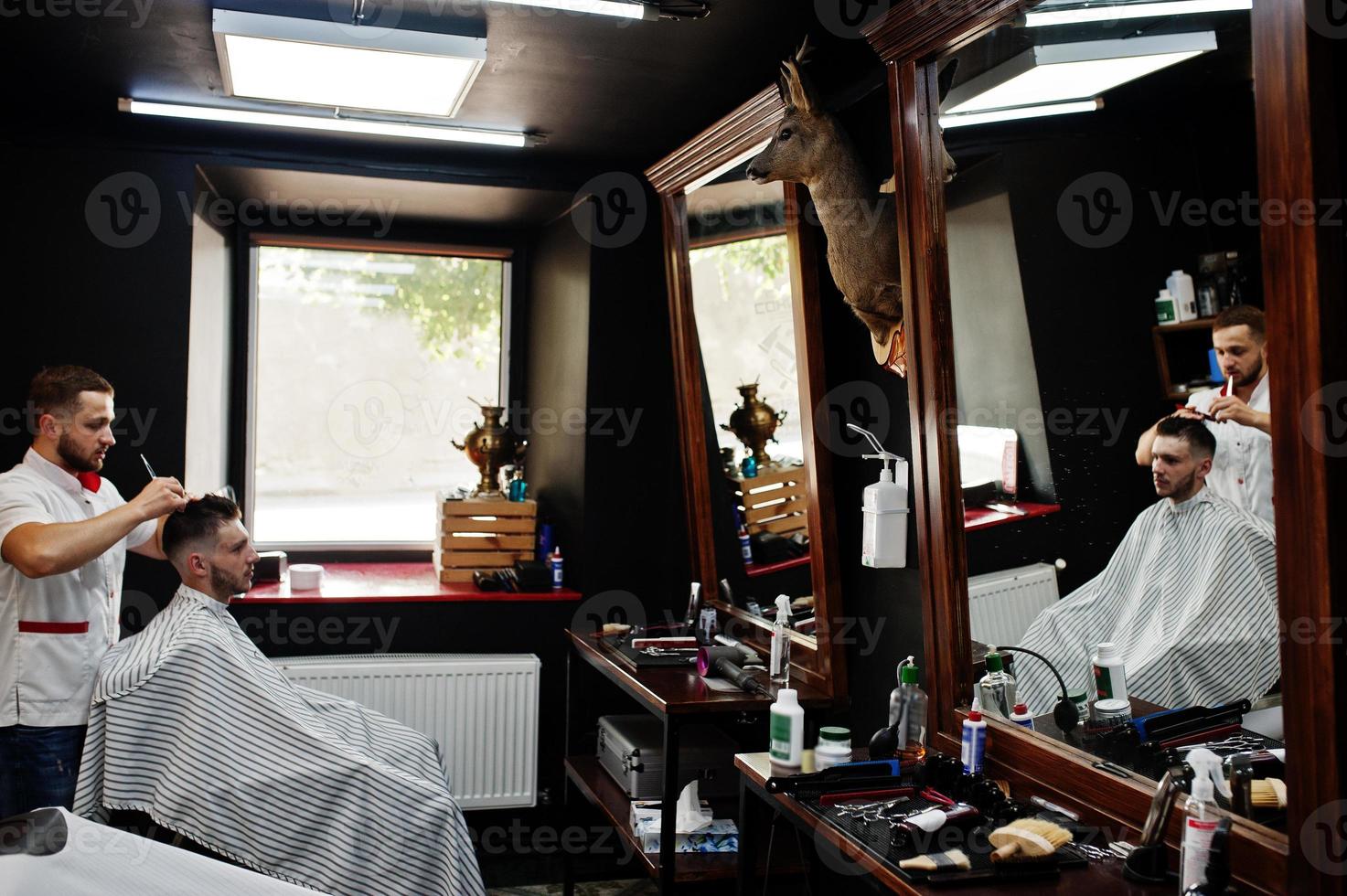 Young bearded man getting haircut by hairdresser while sitting in chair at barbershop. Barber soul. photo