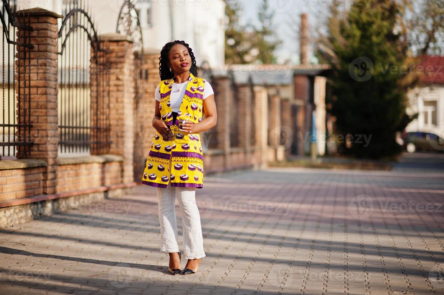 Stylish african american women in yellow jacket posed on street with hot drink in disposable paper cup. photo