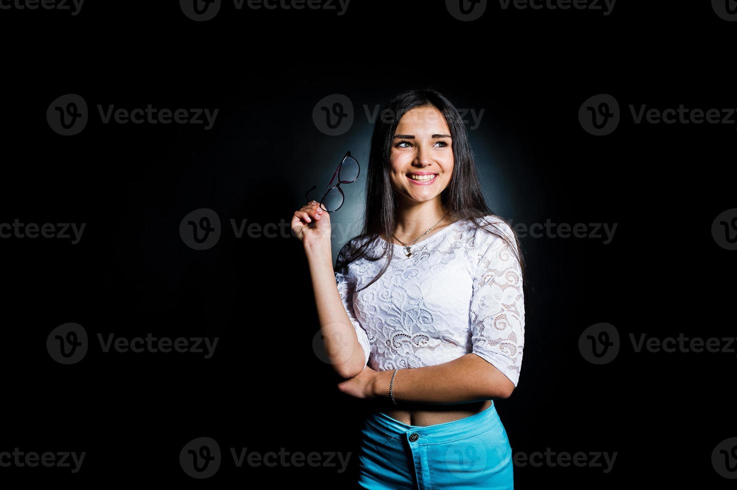 Portrait of an attractive young woman in white top and blue pants posing with her glasses in the dark. photo