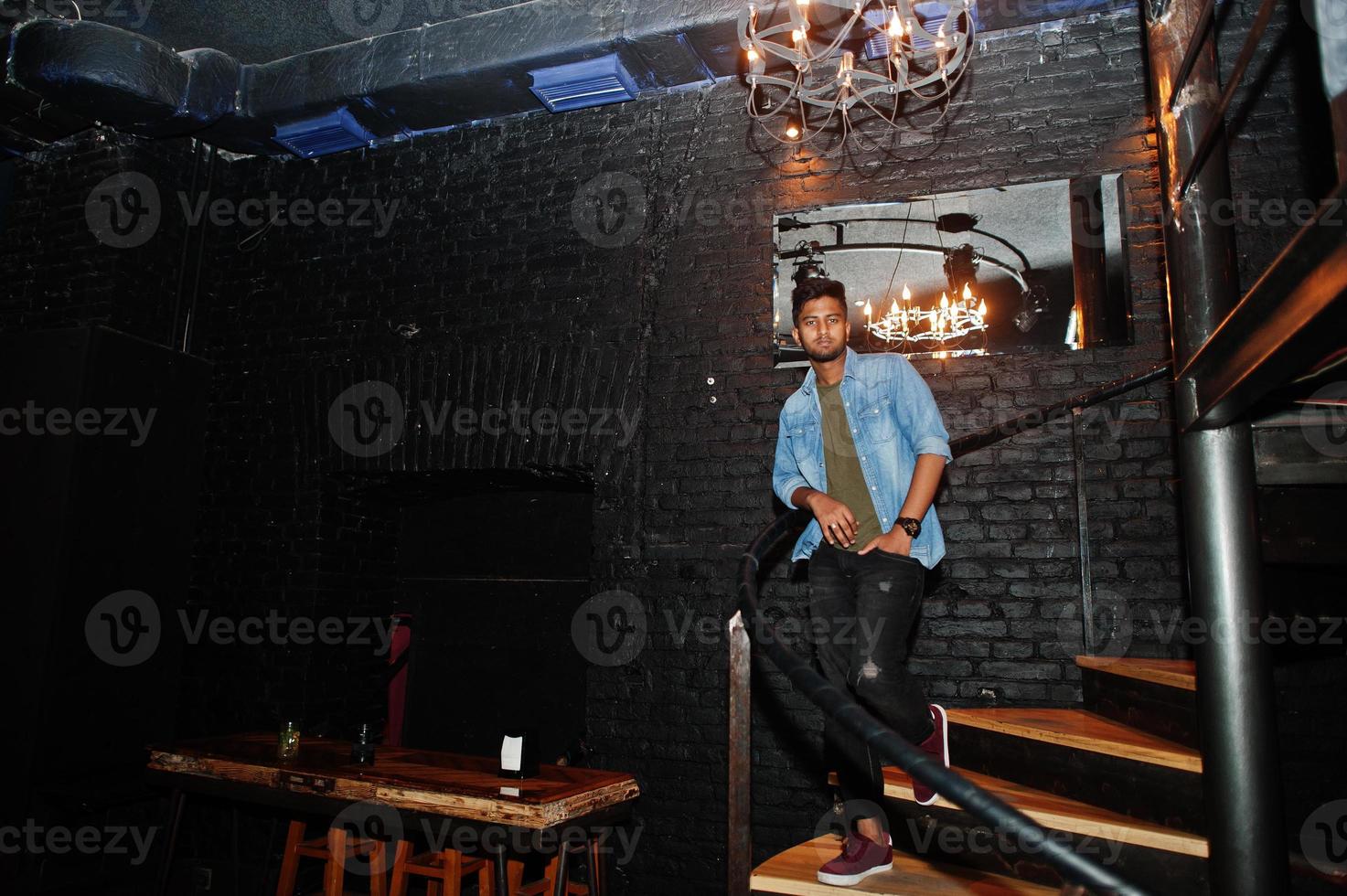 retrato de un apuesto y exitoso sur asiático barbudo, joven autónomo indio con camisa de jeans azul parado en un club nocturno en las escaleras. foto
