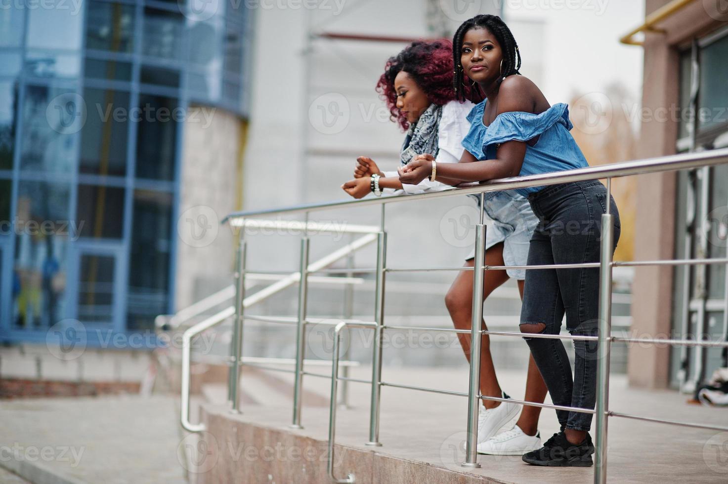 dos atractivas mujeres afroamericanas posaron cerca de las barandillas contra un moderno edificio de varios pisos. foto