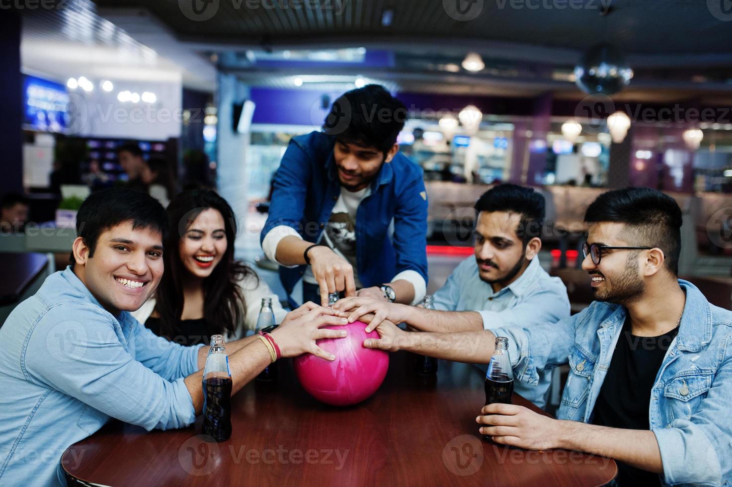 Group of five south asian peoples having rest and fun at bowling club, sitting on table and drinking soda drinks at glass bottles. photo