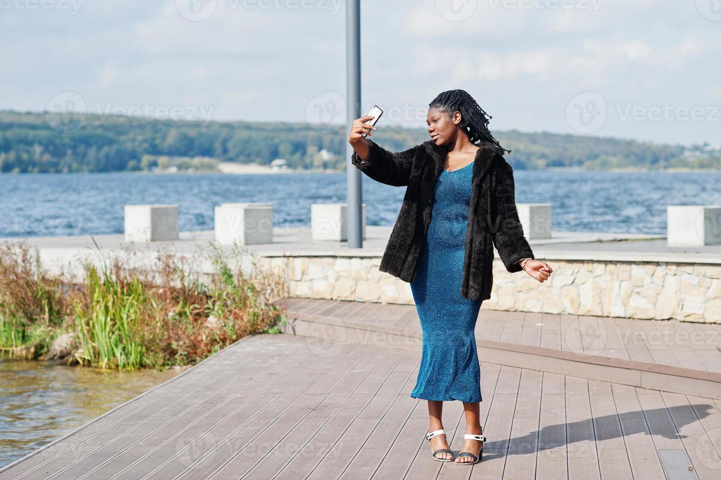 African american dark skinned plus size model posed in a blue shiny dress and black fur coat against sea side making selfie on telephone. photo