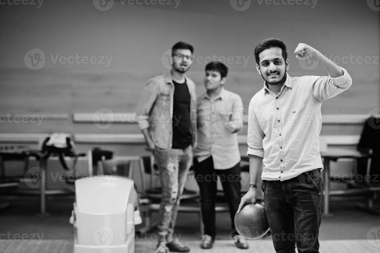 South asian man in jeans shirt standing at bowling alley with ball on hands. Guy is preparing for a throw. Friends support him loudly. photo