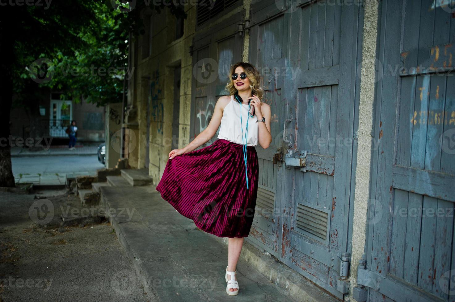 Chica modelo rubia de moda y hermosa con elegante falda de terciopelo rojo, blusa blanca y gafas de sol, posada con teléfono y auriculares. foto