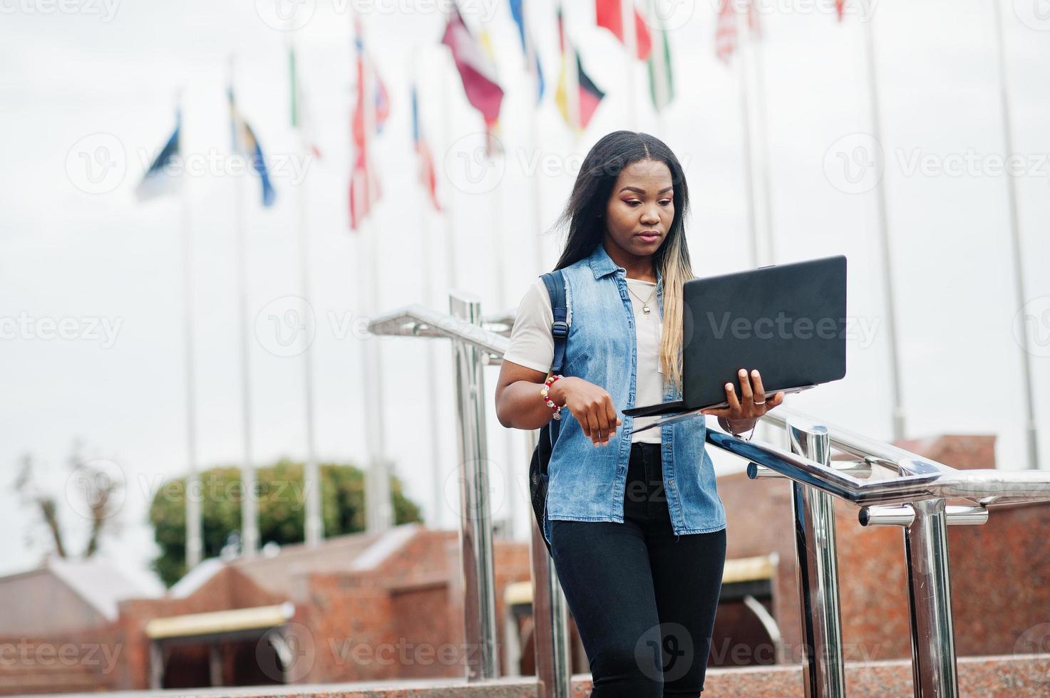 African student female posed with backpack and school items on yard of university, against flags of different countries. She hold laptop on hands. photo