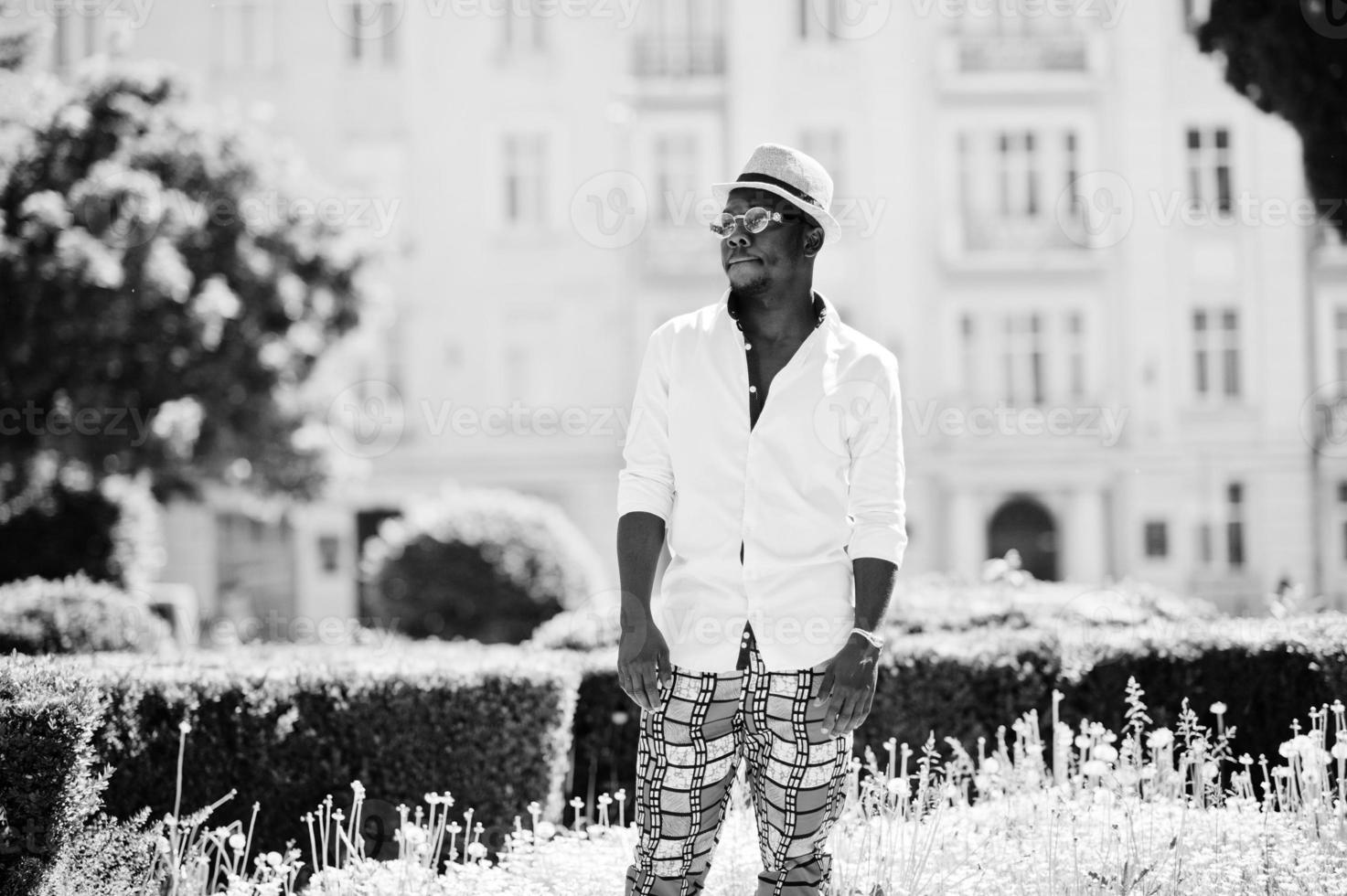 elegante hombre afroamericano con camisa blanca y pantalones de colores con sombrero y gafas al aire libre. chico modelo de moda negro. foto