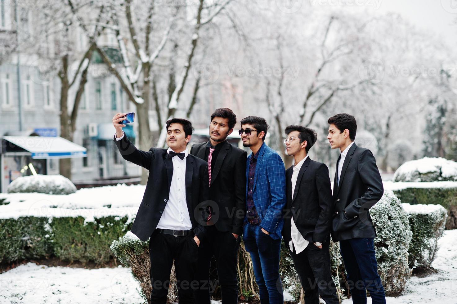 Group of 5 indian students in suits posed outdoor in winter day and making selfie on phone. photo