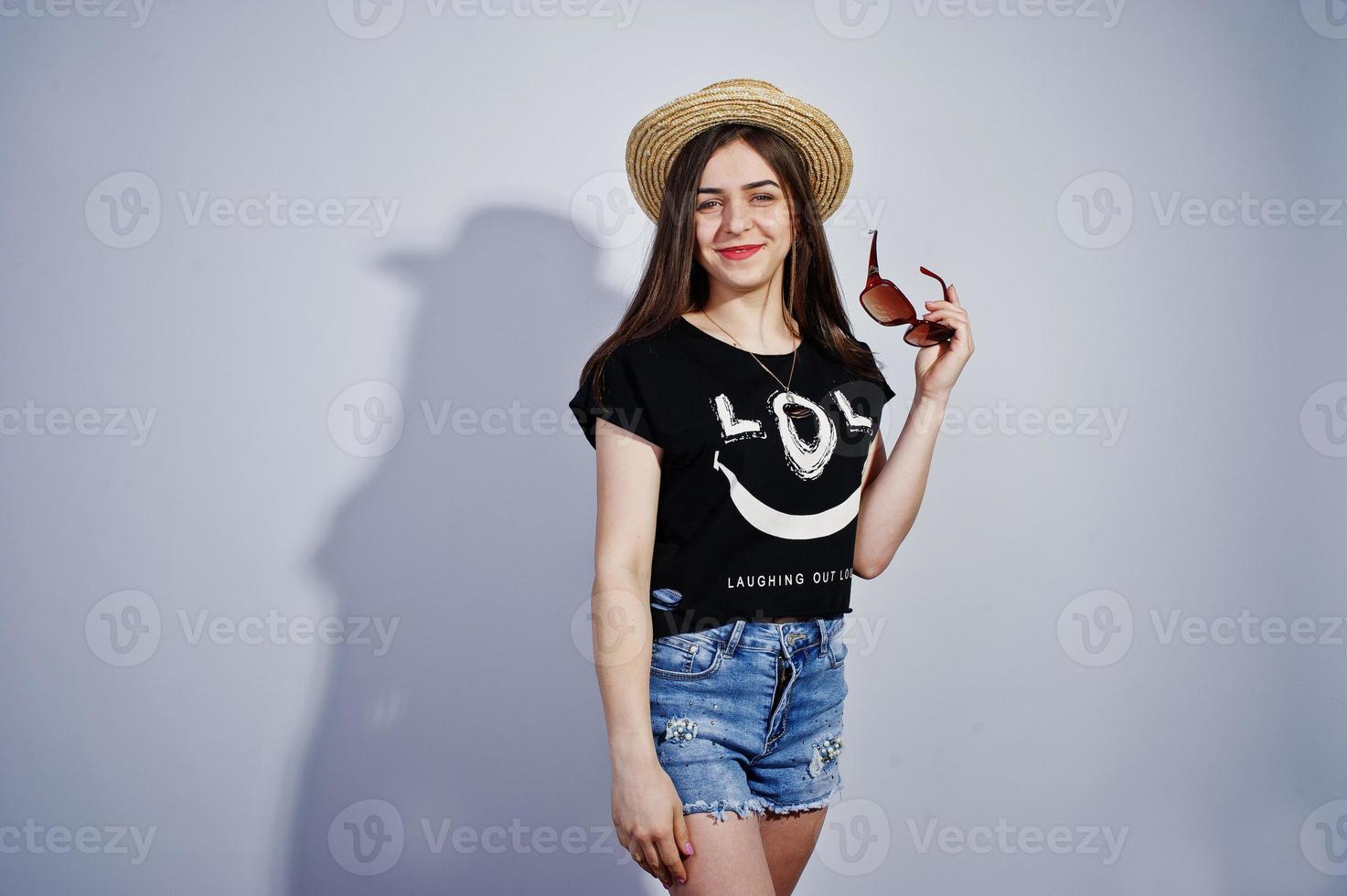 retrato de una chica atractiva en camiseta negra diciendo jajaja, pantalones cortos de mezclilla, sombrero y gafas de sol posando en el estudio. foto