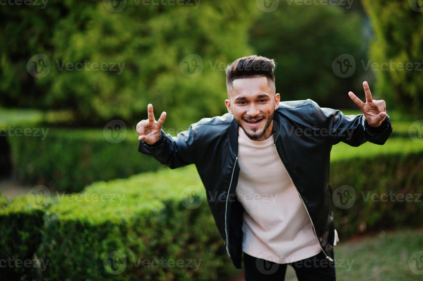Stylish indian beard man at black leather jacket shows two fingers. India model posed at green garden of city. photo