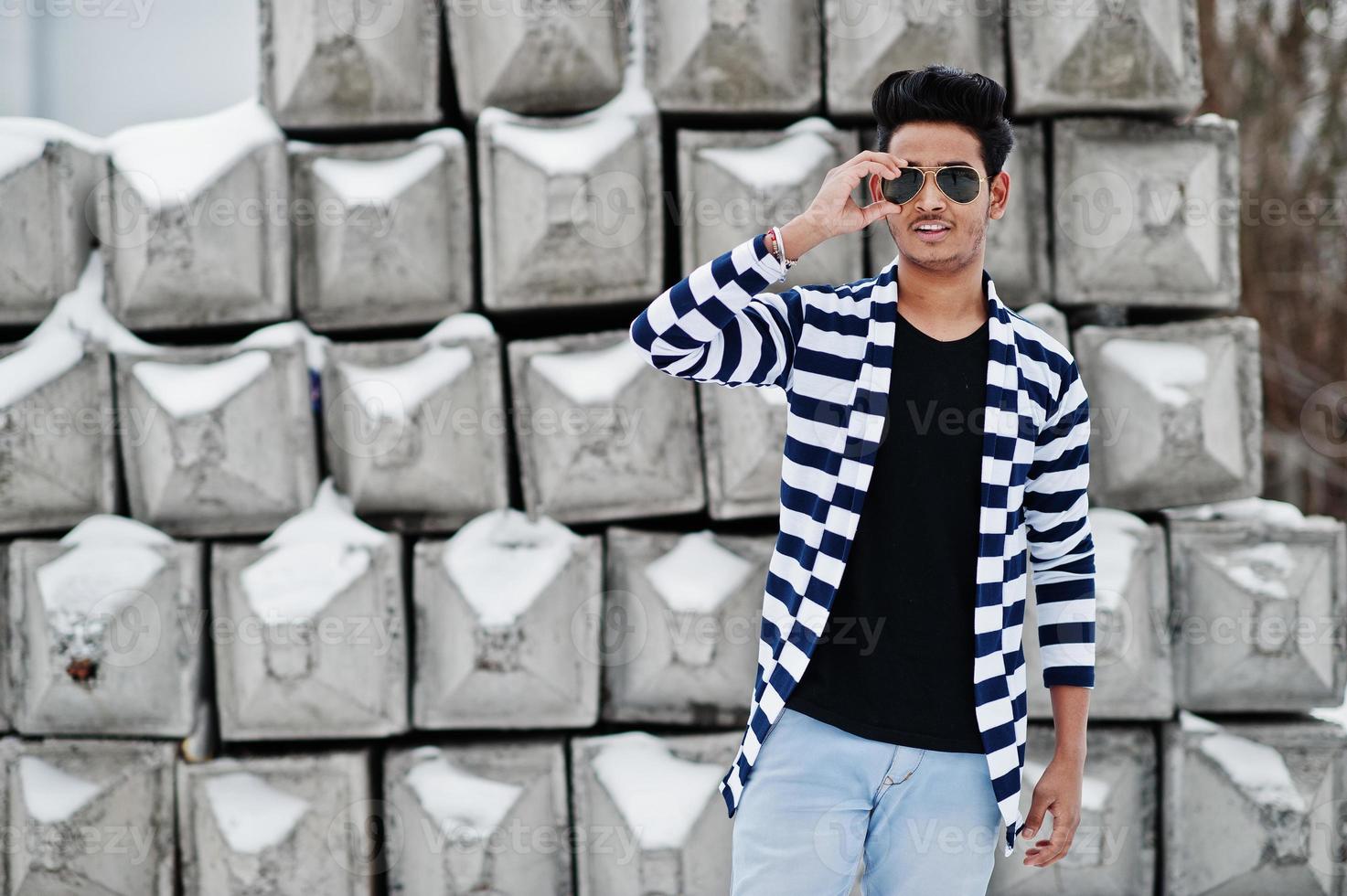 Un joven indio casual con suéter despojado y gafas de sol posó contra bloques de piedra. foto