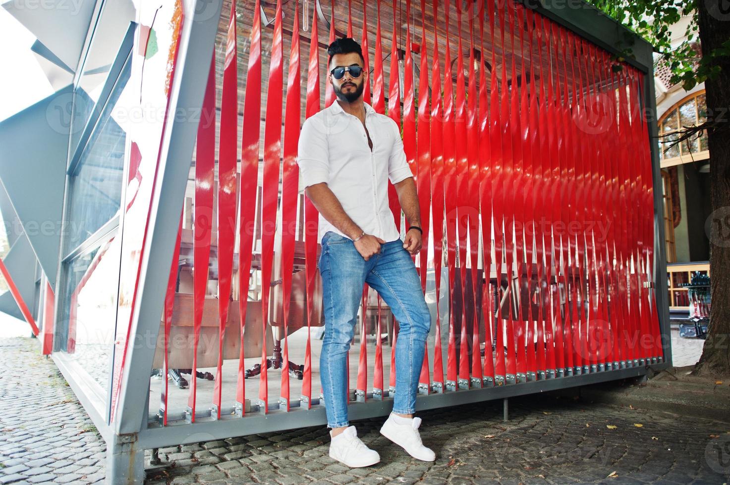 Stylish tall arabian man model in white shirt, jeans and sunglasses posed at street of city. Beard attractive arab guy against red wall. photo