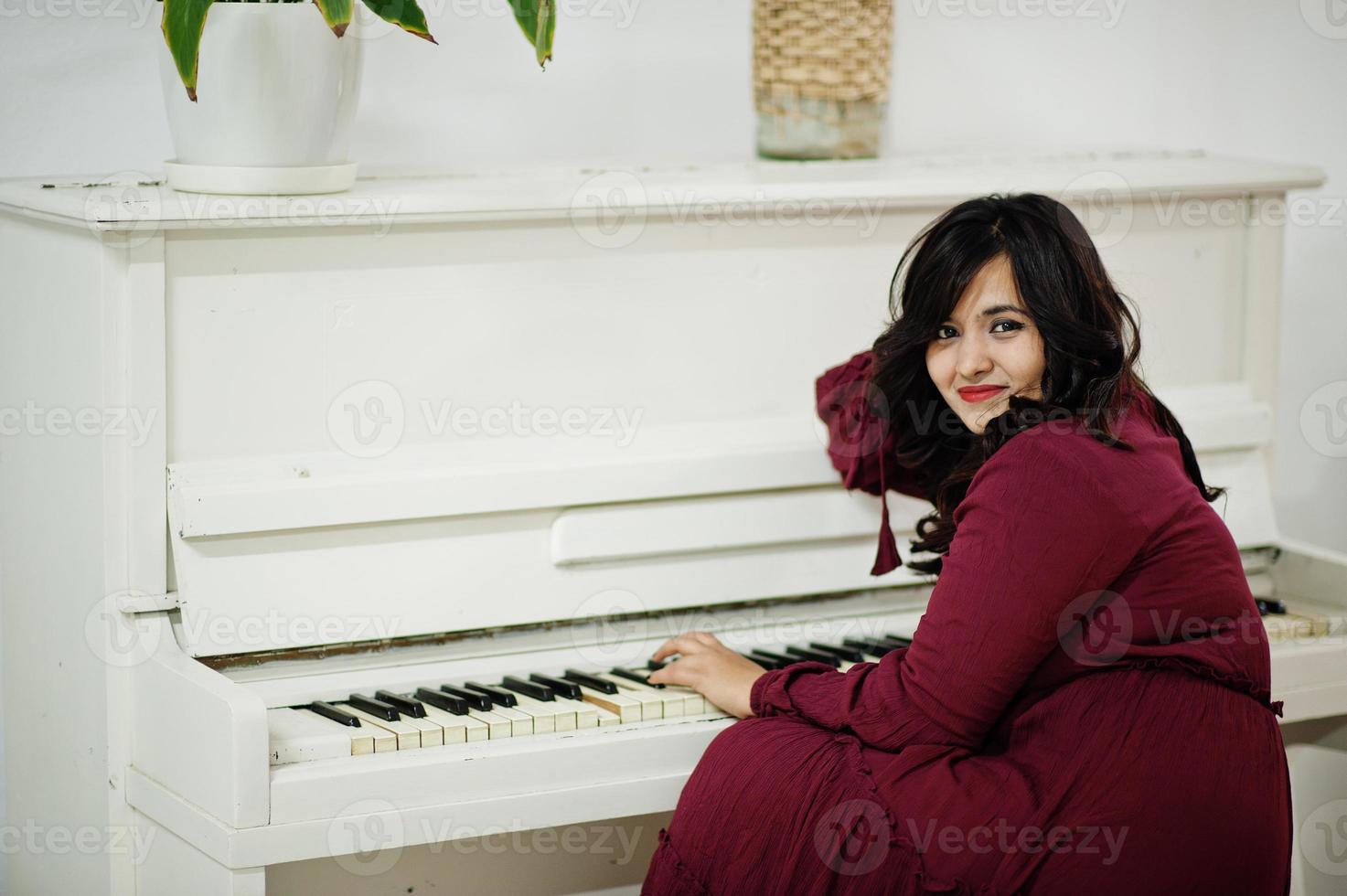 Attractive south asian woman in deep red gown dress posed at room and play on piano. photo
