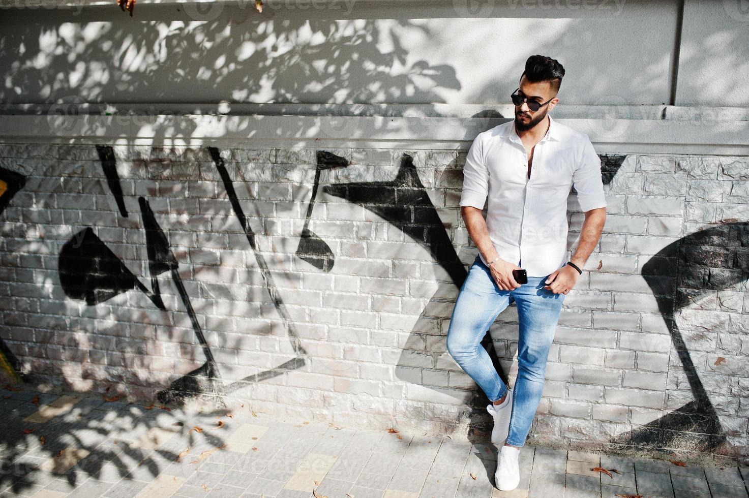 Stylish tall arabian man model in white shirt, jeans and sunglasses posed at street of city. Beard attractive arab guy against graffiti wall. photo