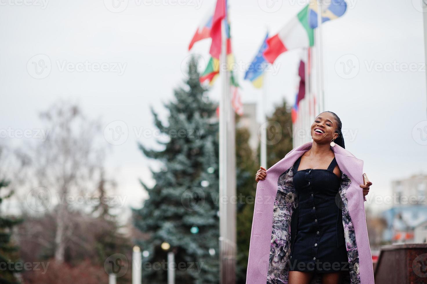 Young stylish beautiful african american woman in street, wearing fashion outfit coat, against flags of different countries of the world. photo