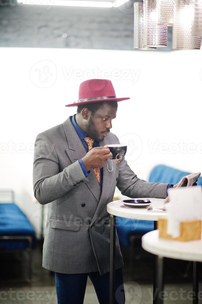 Stylish African American man model in gray jacket tie and red hat drink coffee at cafe and read newspapers. photo