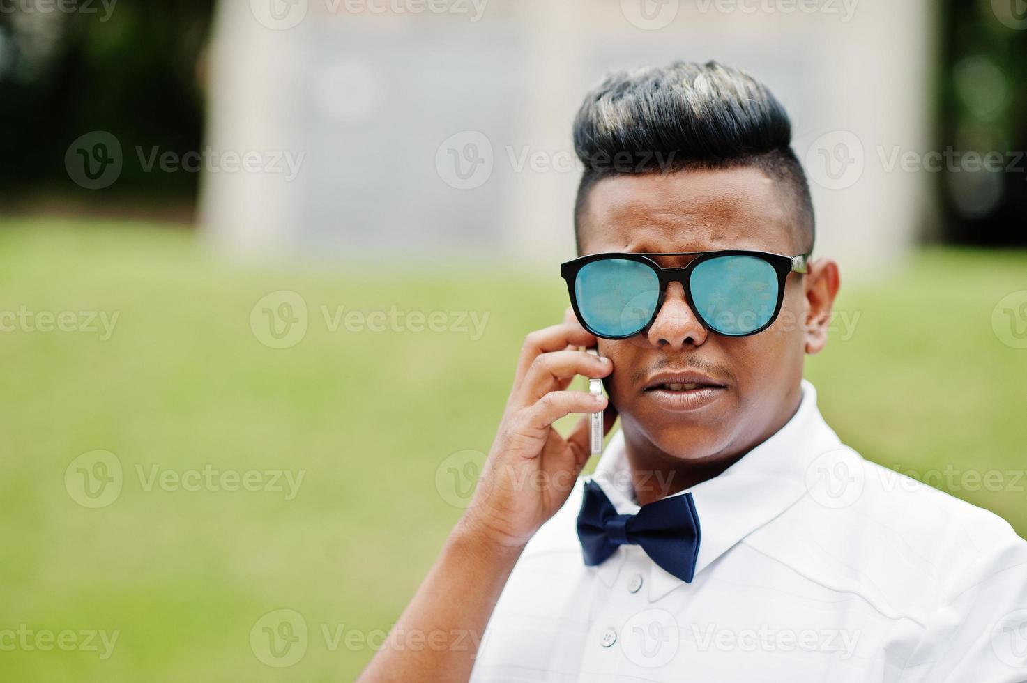 Close up portrait of stylish arabian man at sunglasses and bow tie posed outdoor and speaking on mobile phone. Arab model boy. photo