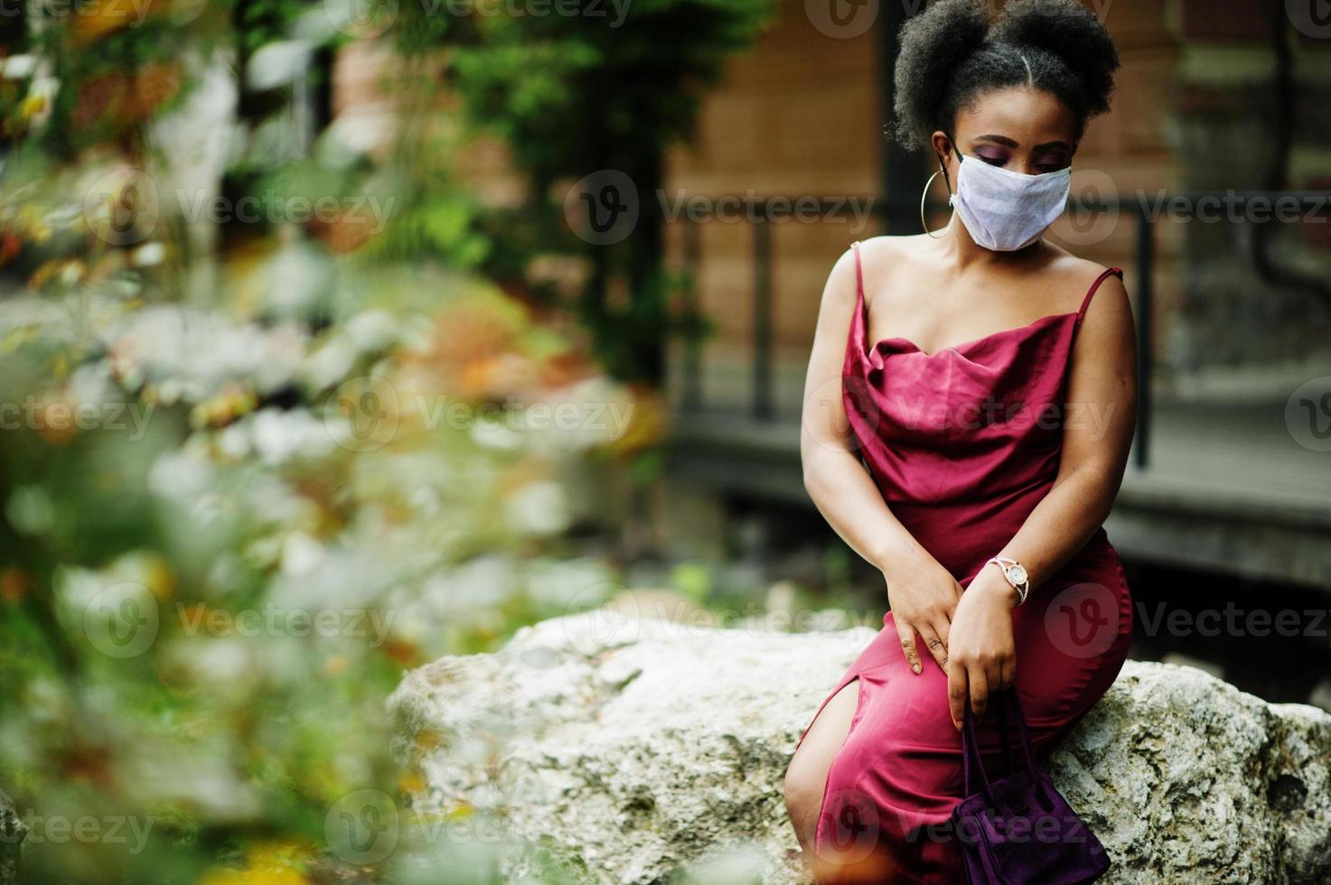 Covid-19, infectious virus. African woman with curly hair, wears red silk dress and medical disposable mask, cares about her health and protects in dangerious situation. photo