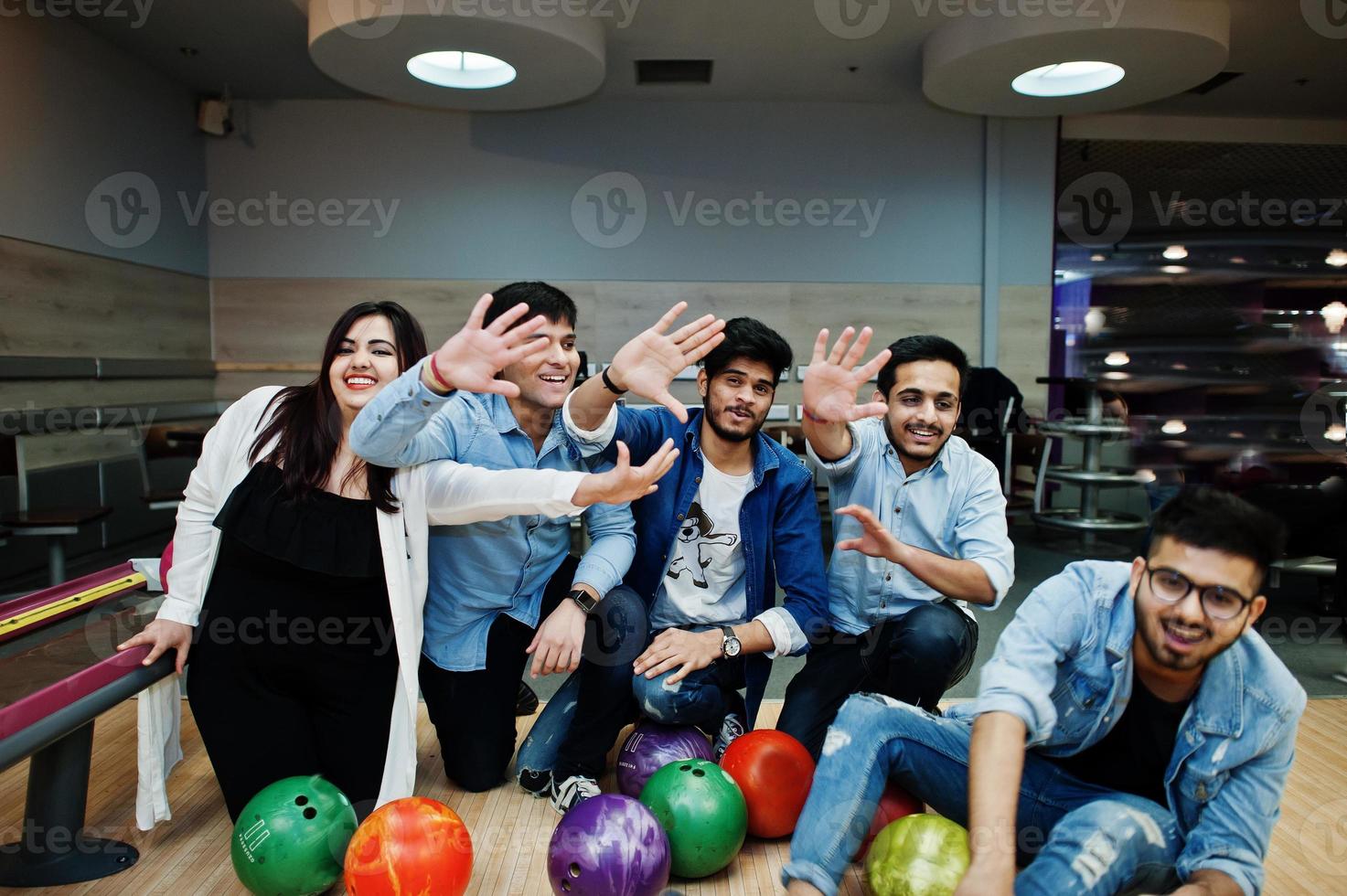 Group of five south asian peoples having rest and fun at bowling club. Holding bowling balls, sitting on alley and giving high five by hands. photo