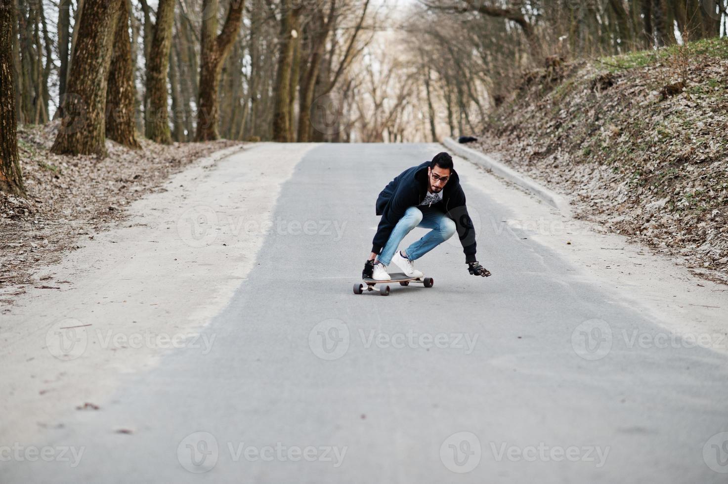 Street style arab man in eyeglasses with longboard longboarding down the road. photo