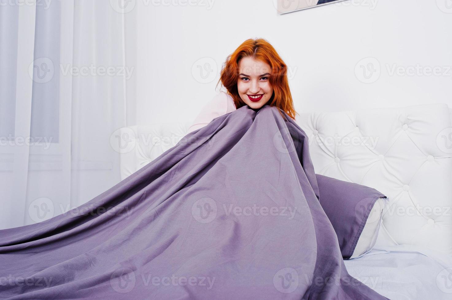 Portrait of an attractive redheaded girl laying on the bed and covering herself with a purple blanket. photo