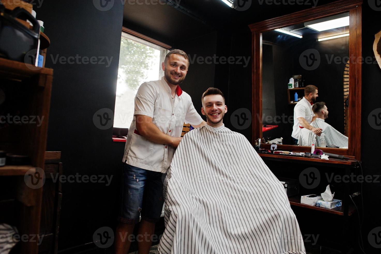 Young bearded man getting haircut by hairdresser while sitting in chair at barbershop. Barber soul. photo