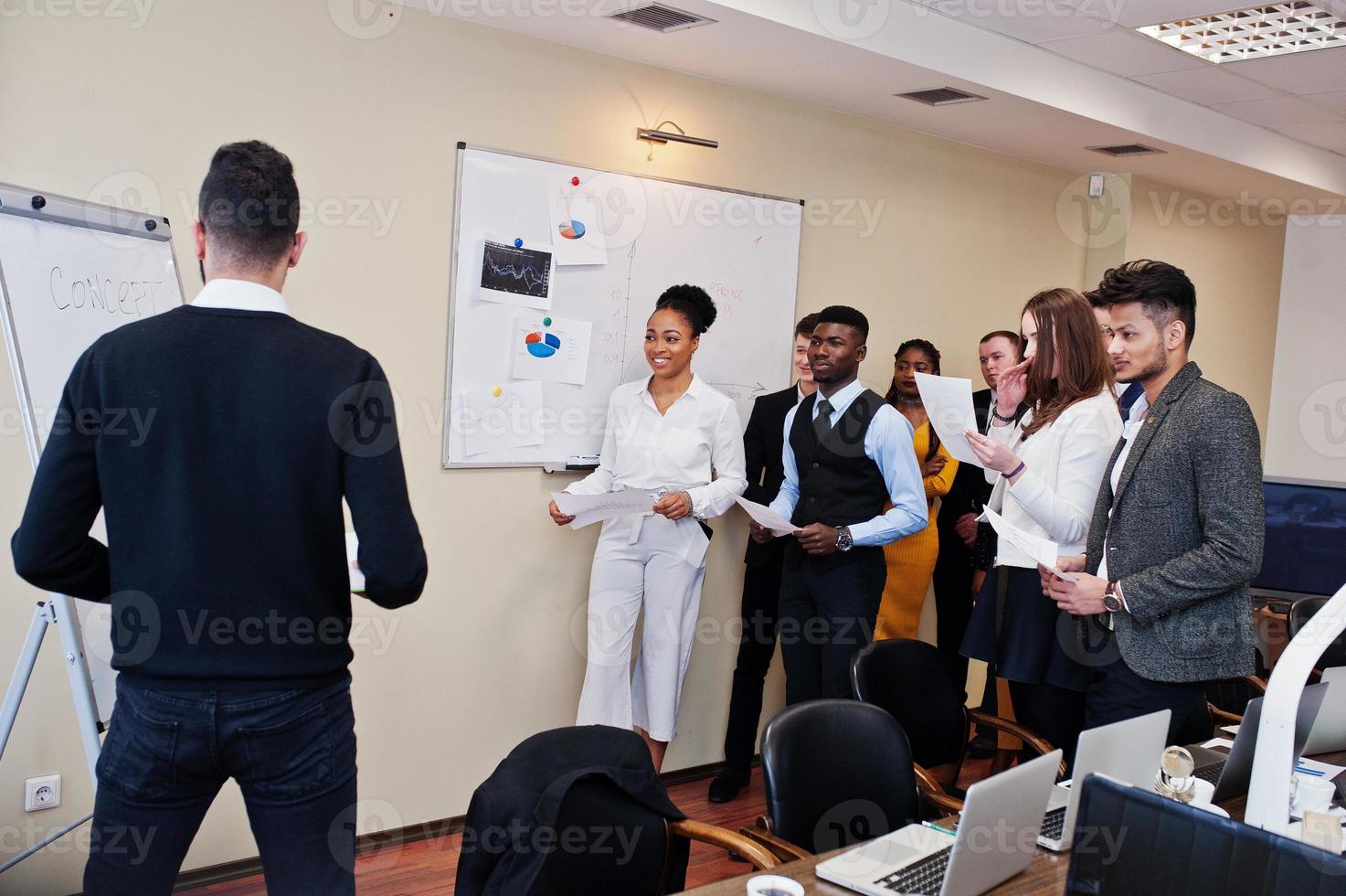 Mixed race business coach presenting report standing near whiteboard pointing on sales statistic shown on diagram and chart teach diverse company members gathered together in conference room. photo