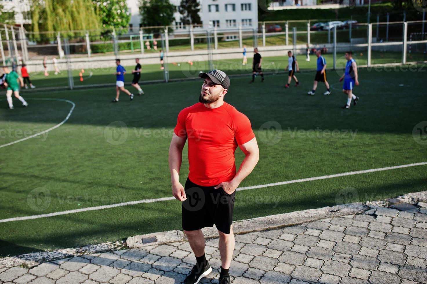 Young brutal bearded muscular man wear on red shirt, shorts and cap at stadium. photo