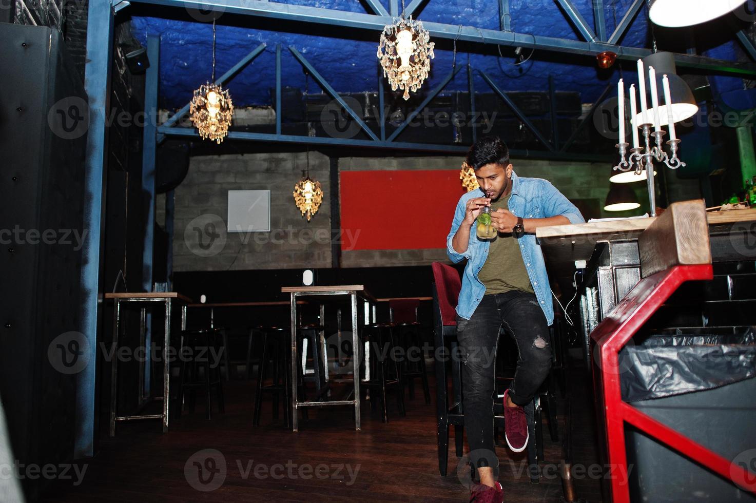 Portrait of handsome successful bearded south asian, young indian freelancer in blue jeans shirt sitting in night club against bar counter with cocktail and having a rest. photo