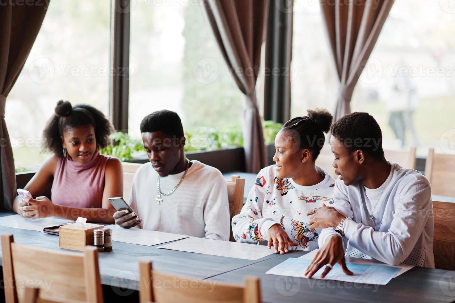 Happy african friends sitting and chatting in cafe. Group of black peoples meeting in restaurant and look at their mobile phone. photo