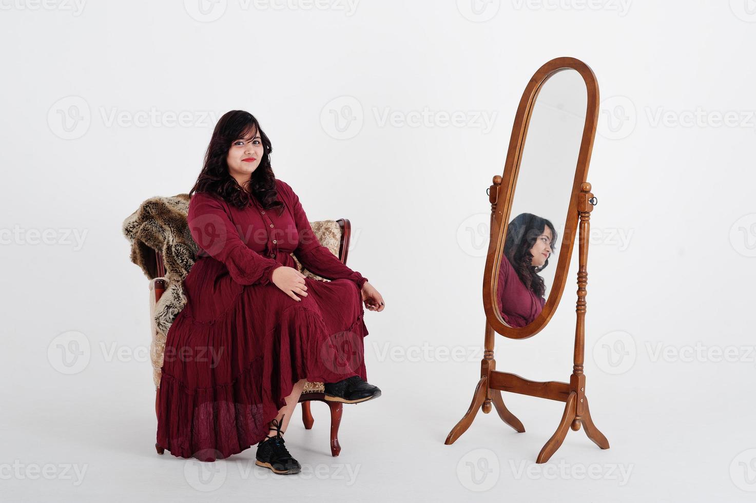 Attractive south asian woman in deep red gown dress posed at studio on white background against mirror and sitting at chair. photo