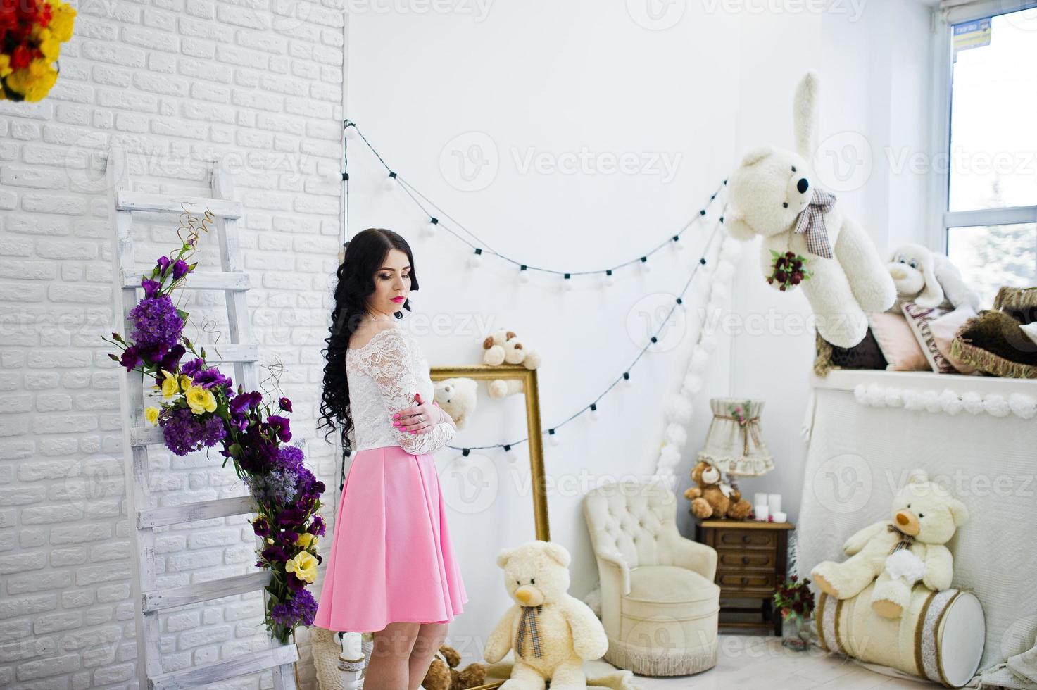 Young brunette girl in pink skirt and white blouse posed indoor against room with toys bear. photo