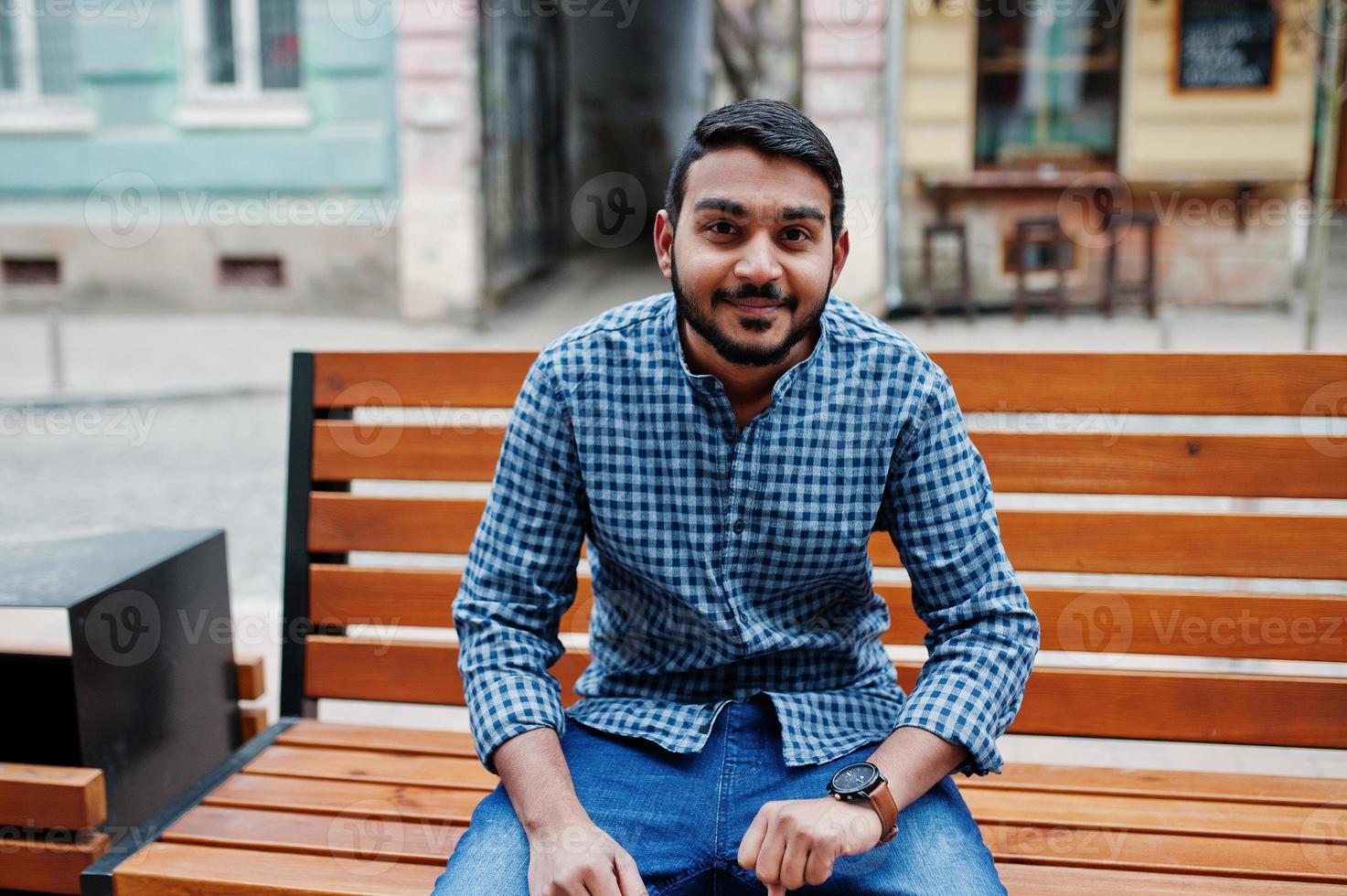 Stylish indian beard model man in casual clothes posed outdoor, sitting on bench at street of India and looking at his watches. photo