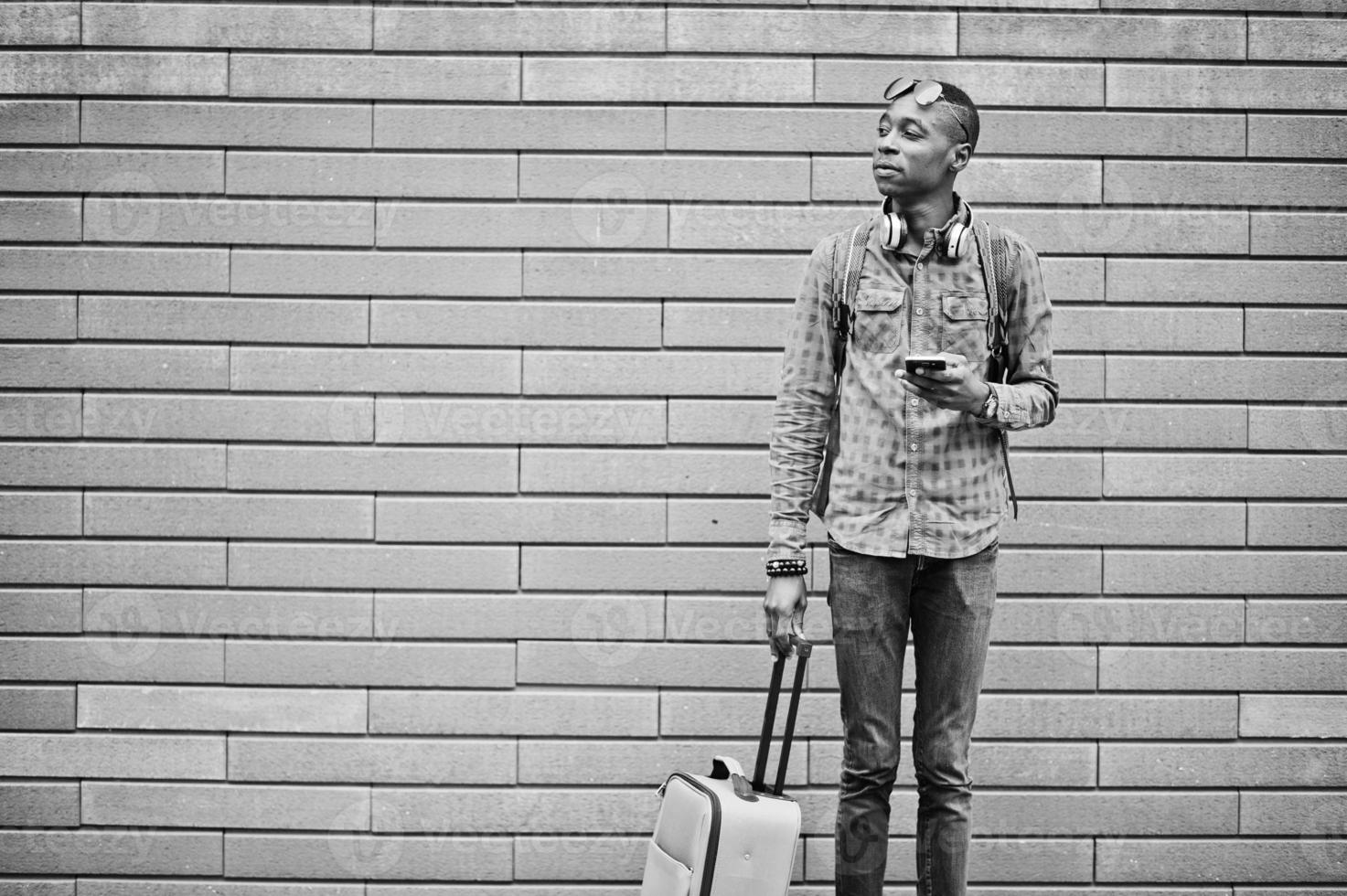 African american man in checkered shirt, sunglasses and earphones with suitcase and backpack. Black man traveler against wall holding mobile phone. photo