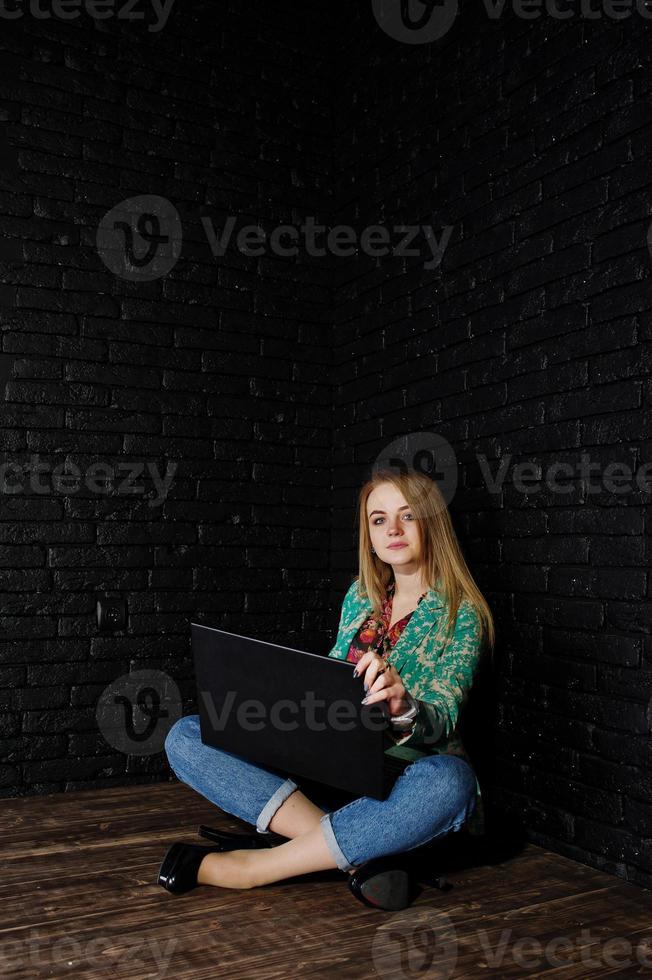 Stylish blonde girl in jacket and jeans with laptop against brick black wall at studio. photo