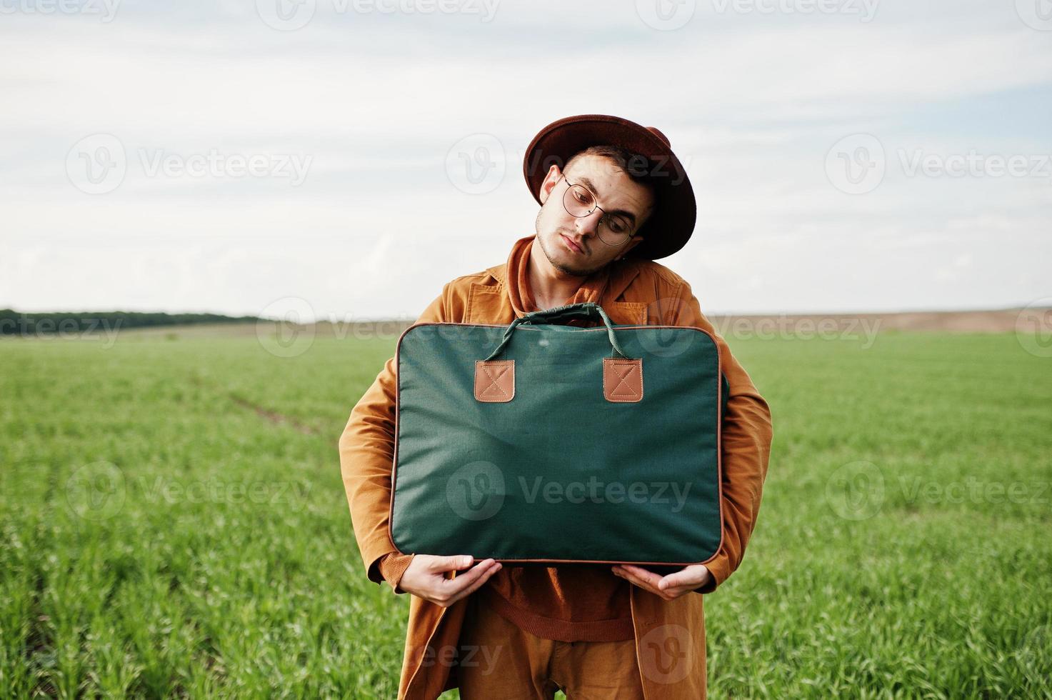 Stylish man in glasses, brown jacket and hat with bag posed on green field. photo