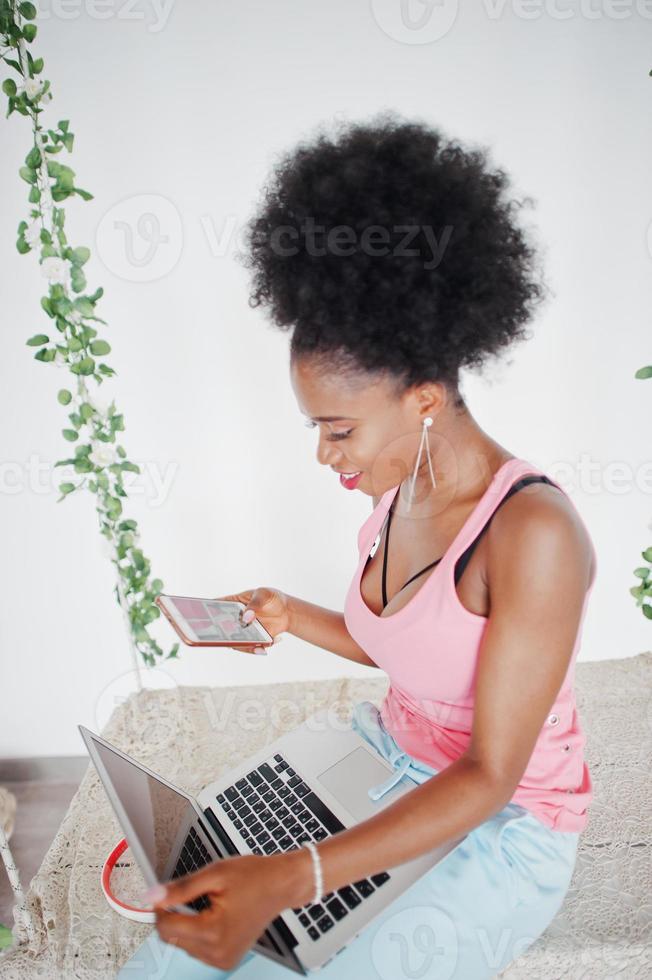 Young african american woman sitting in swing while working on laptop and looking at mobile phone. photo