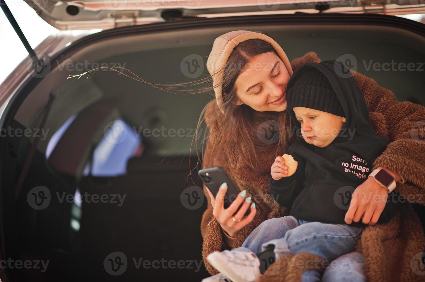 Young mother and child sitting in the trunk of a car and looking at mobile phone. Safety driving concept. photo