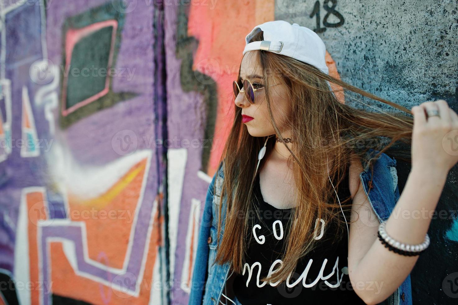 elegante chica hipster casual con gorra, gafas de sol y jeans, escuchando música desde auriculares de teléfono móvil contra una gran pared de graffiti. foto