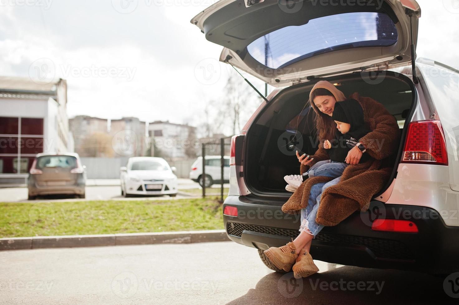 Young mother and child sitting in the trunk of a car and looking at mobile phone. Safety driving concept. photo