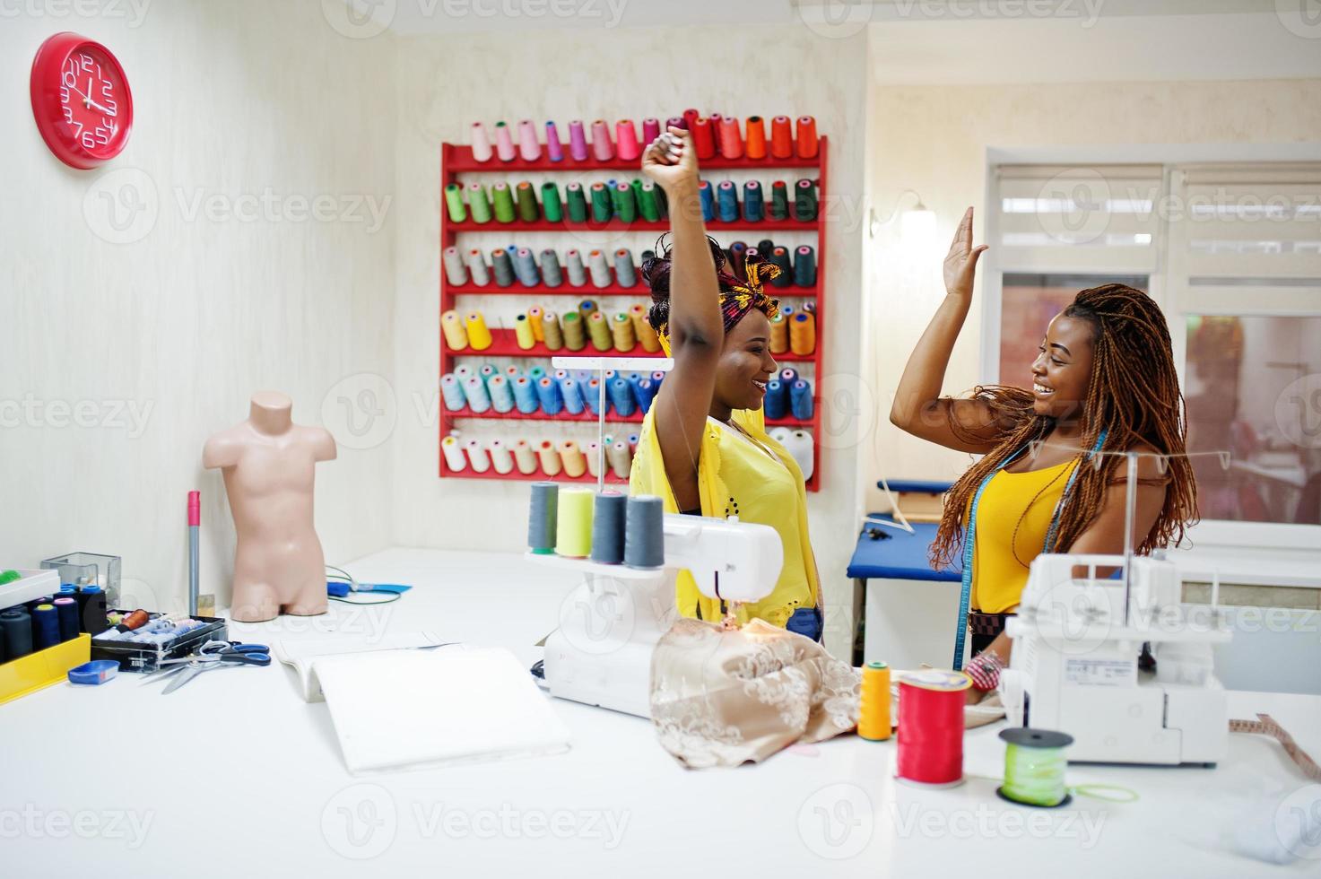 Two african dressmaker woman sews clothes on sewing machine at tailor office. Black seamstress girls gives high five each other. photo